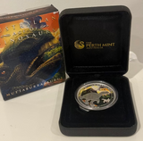 Silver coin in a black case with 'The Perth Mint' logo, next to a dinosaur-themed book on a white background.